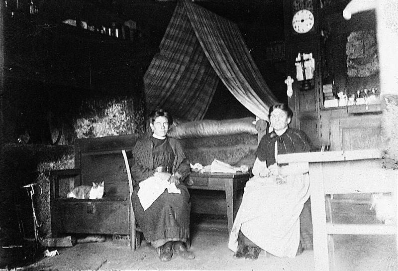 Deux femmes cousant ou tricotant devant un coin de feu (canton), un lit (lièch) et une pendule (relòtge), en Aubrac (secteur de Laguiole)