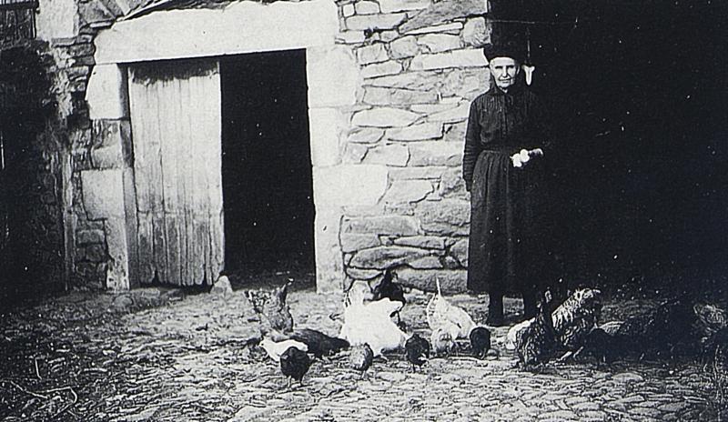  Femme ayant levé les œufs (uòus) et poules (galinas, polas) devant une porte d'étable, en Aubrac (secteur de Saint-Chély d’Aubrac), vers 1930