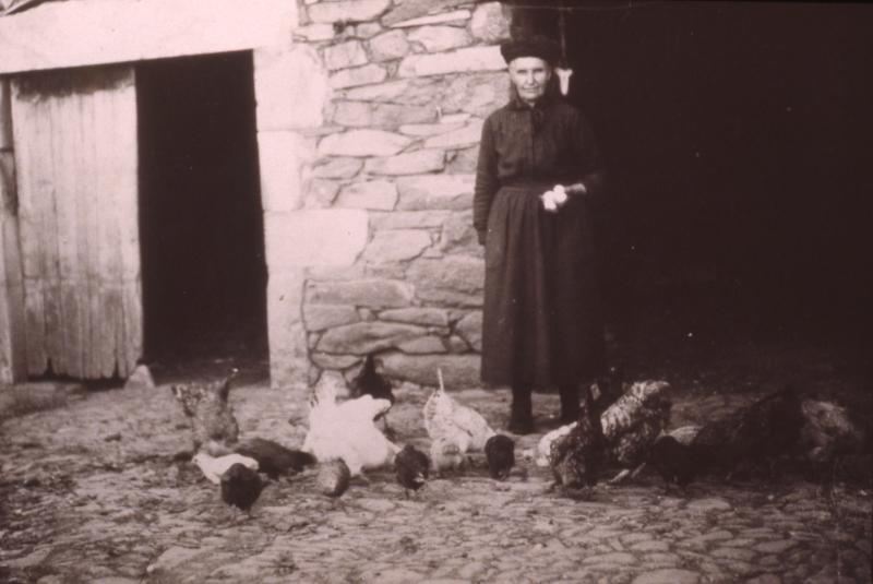  Femme ayant levé les œufs (uòus) et poules (galinas, polas) devant une porte d'étable, en Aubrac (secteur de Saint-Chély d’Aubrac), vers 1930