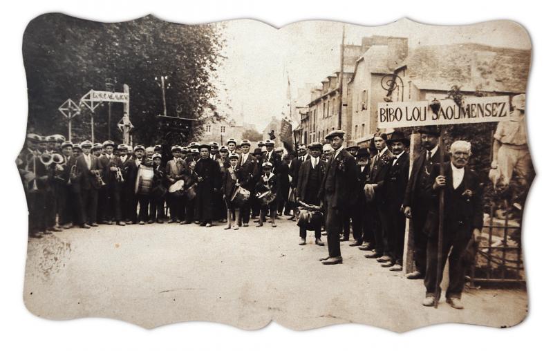 Lyre et villageois (vilatjors) avec pancarte “Bibo lou saoumensez” (Viva lo saumancés) rassemblés au monument aux morts pour la Saint-Bourrou, années 1930