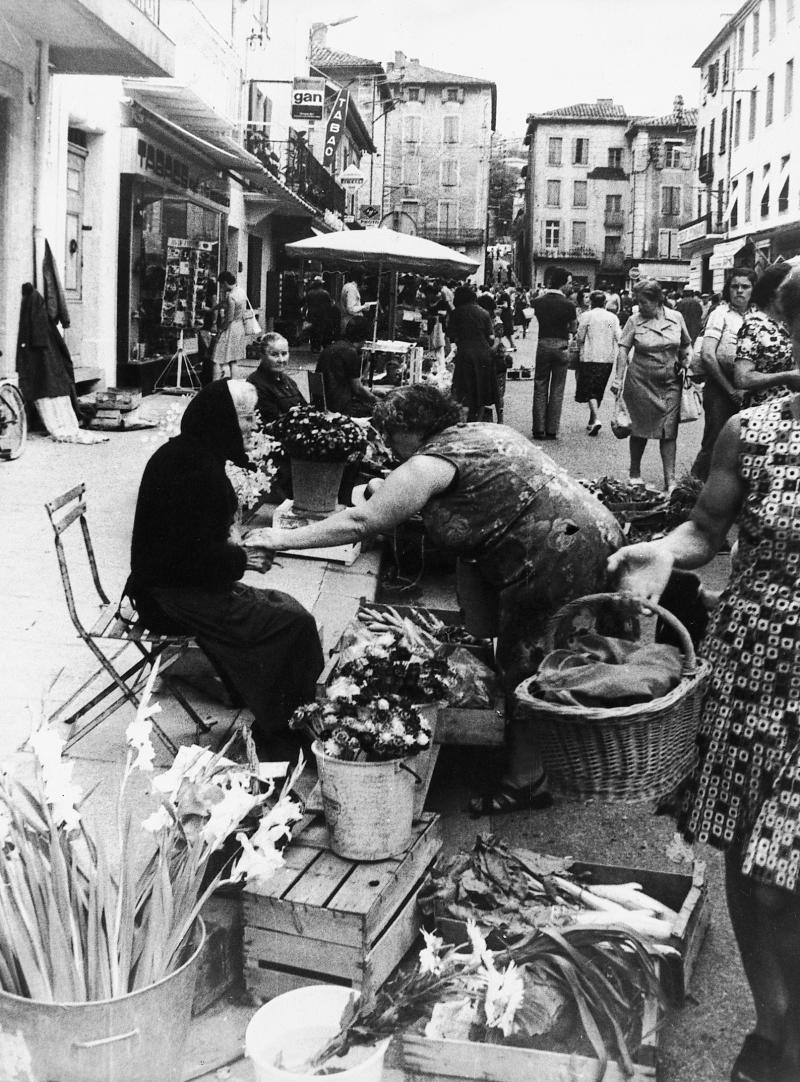  Etal d'une marchande (merchanda) de fleurs et légumes et clientes dans une rue (carrièira) un jour de foire (fièira) ou de marché (mercat), 1976
