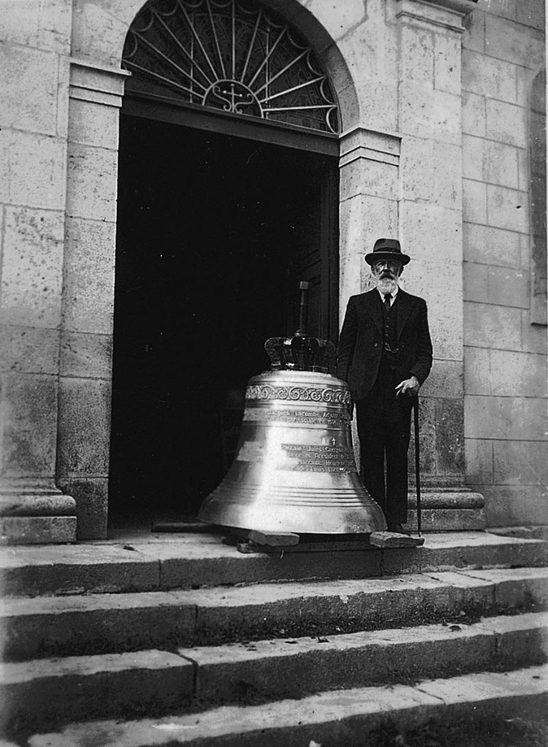 Homme posant à côté d'une cloche (campana) devant la porte d'entrée de l'église (glèisa), à Frons, 1932