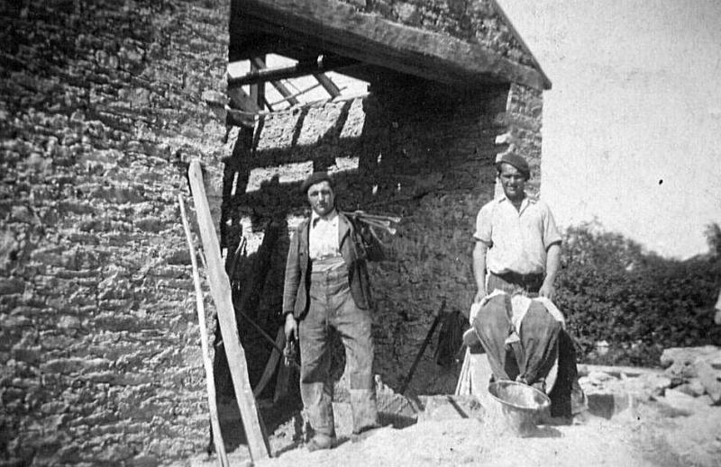 Deux maçons (peirièrs) ou ouvriers (obrièrs) sur un tas de pierres devant une grange (fenial, granja) en construction
