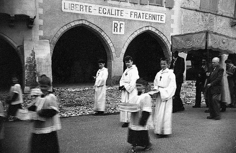 Enfants de chœur (clergues) et marguilliers (marguilhièrs) portant le dais devant la mairie (comuna, ostal comuna) durant une procession de la Fête Dieu (Fèsta Diu), 1954