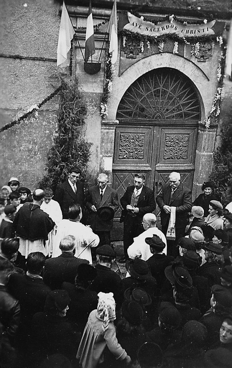 Réception par les fabriciens (fabricians) du nouveau curé (curat) devant la porte de l'église (glèisa) pavoisé, 1957