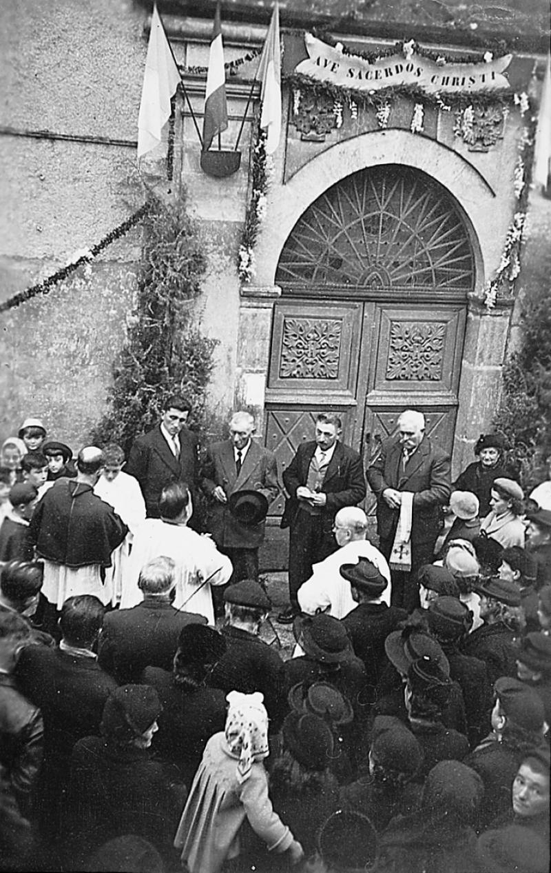 Réception par les fabriciens (fabricians) du nouveau curé (curat) devant la porte de l'église (glèisa) pavoisé, 1957