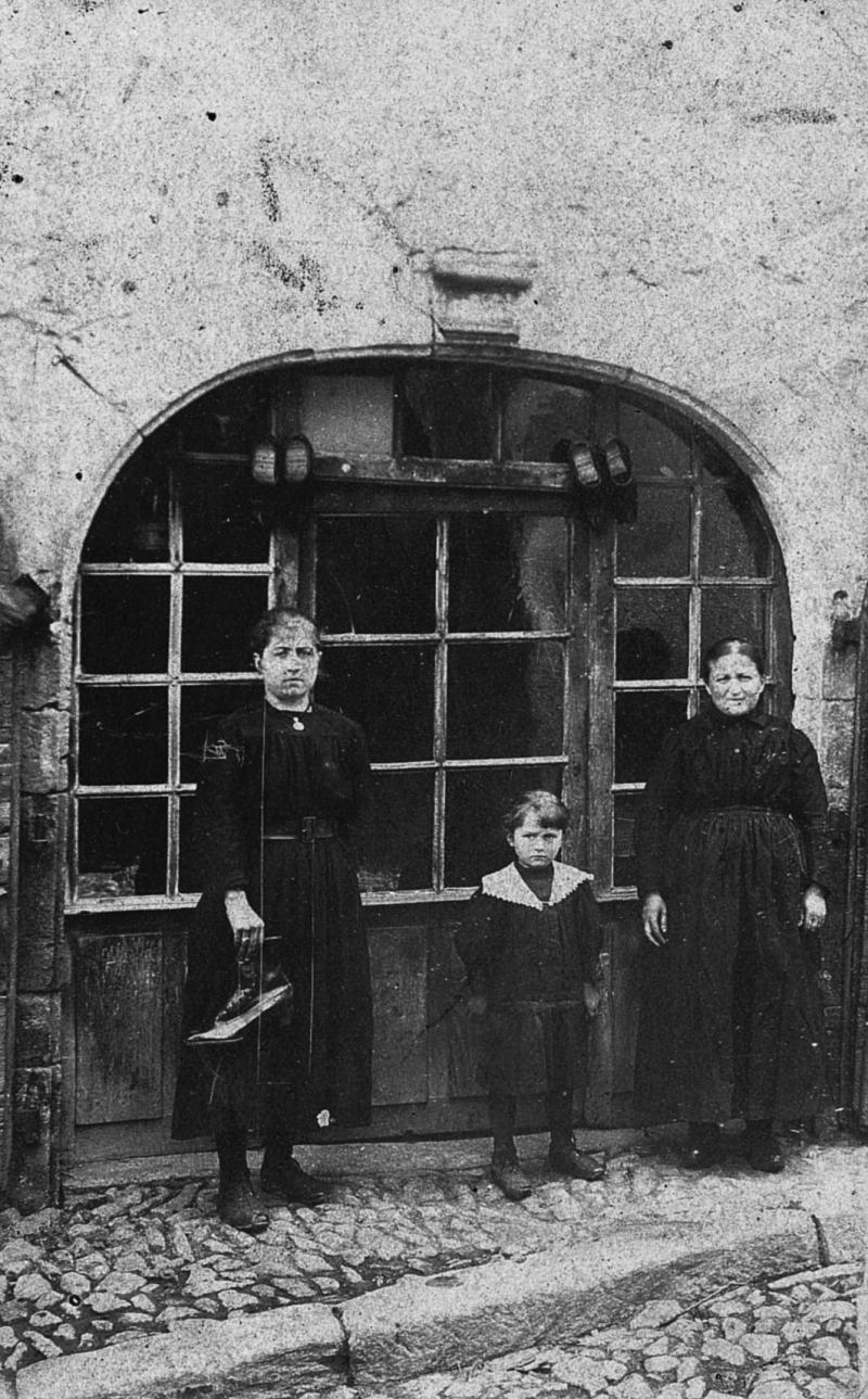 Deux enfants (dròlles, enfants) avec leur mère (maire) devant la devanture (veirina) d'un sabotier (esclopièr), rue droite, 1917