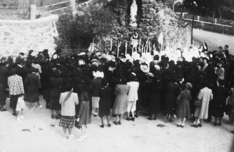 Paroissiens (parroquians), communiantes avec cierge, curé (curat) et religieuses (sòrres, surs) rassemblés devant une reconstitution de la grotte de Lourdes